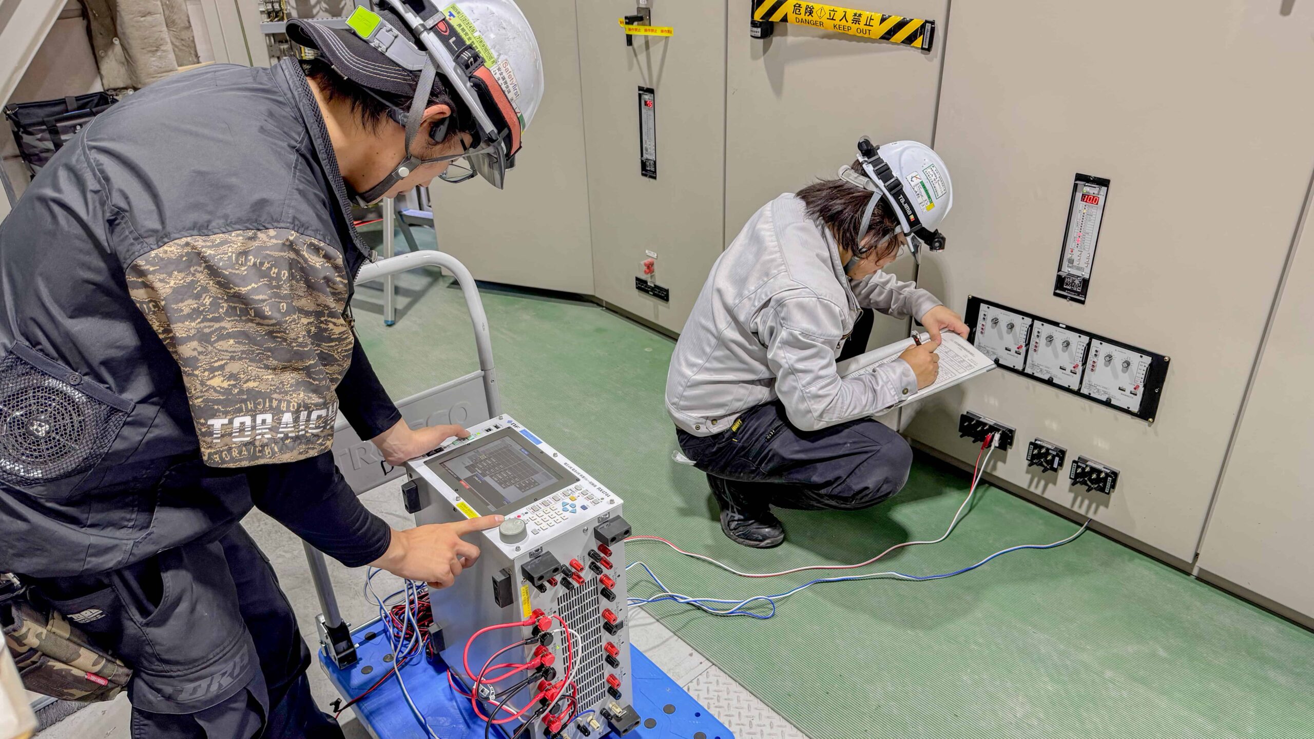 保護継電器試験、機器を繋いで操作する作業員と、チェックする作業員